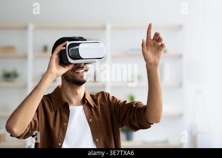Middle Eastern man wearing VR glasses experiencing virtual reality gesturing hand while standing at home Stock Photo