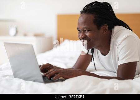 Cheerful Black man with dreads using laptop lying on bed at home Stock Photo