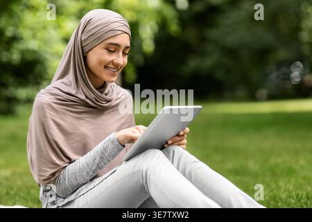 Arab woman wearing hijab using tablet while sitting in park during daytime Stock Photo