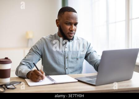 Serious Black businessman working on laptop and holding stylus pen while sitting at table with journal at home Stock Photo