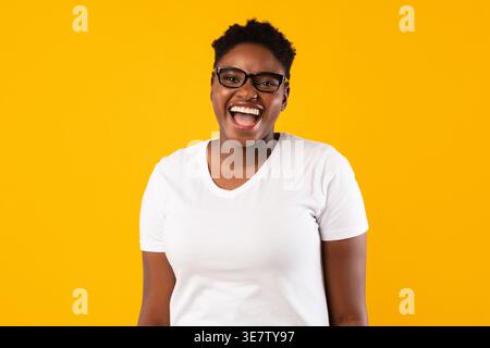Cheerful Black woman wearing eyeglasses standing against yellow background in studio Stock Photo