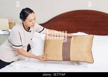 Asian female attendant putting pillows on bed in hotel room Stock Photo