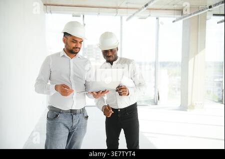 Diverse group of male engineers wearing hard hats holding laptop discussing project during work on construction site Stock Photo