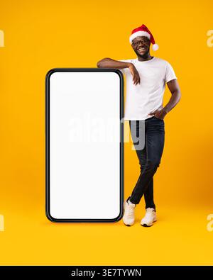African American Guy Standing Near Big Cellphone With Empty Screen Wearing Santa Hat Over Yellow Background. Mobile Application Advertisement, Christm Stock Photo