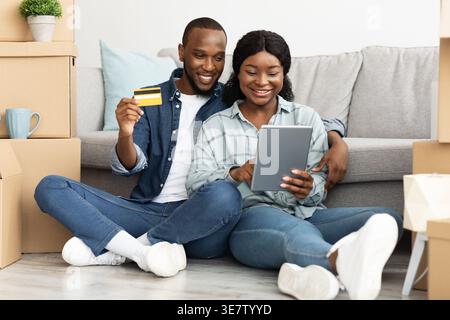 Black husband holding credit card and wife using tablet while sitting together on floor in living room near unpacked boxes at home Stock Photo