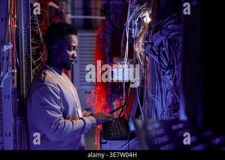 Black male system administrator standing and using computer in server room lit by blue neon light Stock Photo