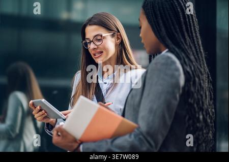 Black businesswoman with braids holding book talking to caucasian female colleague showing tablet standing outside office building during daytime Stock Photo