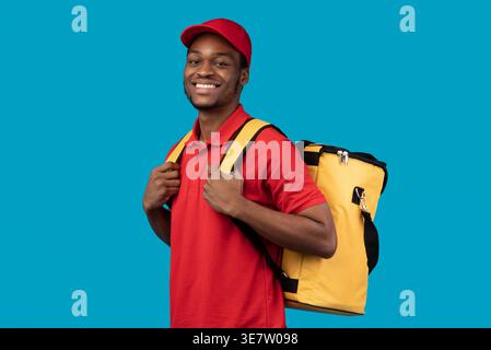 Black delivery man wearing cap carrying thermo backpack showing thumbs up standing against blue background in studio Stock Photo