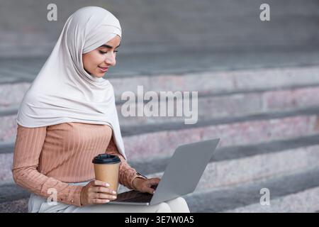 Smiling Indian Muslim woman wearing hijab holding cup of coffee working on laptop sitting on stairs in park during daytime Stock Photo