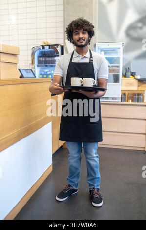 Asian male waiter with curly hair holding tray with cup of coffees while standing in coffee shop Stock Photo