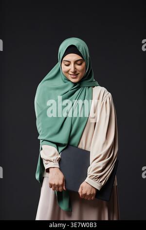Mixed-race woman wearing hijab carrying laptop while standing against black wall in studio Stock Photo