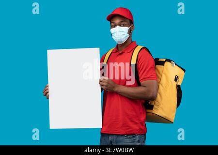 Black delivery man wearing facemask holding blank white board carrying thermo backpack standing against white background in studio Stock Photo