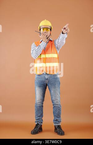 Asian male civil engineer in vest and hardhat pointing away talking on walkie talkie standing against brown background in studio Stock Photo