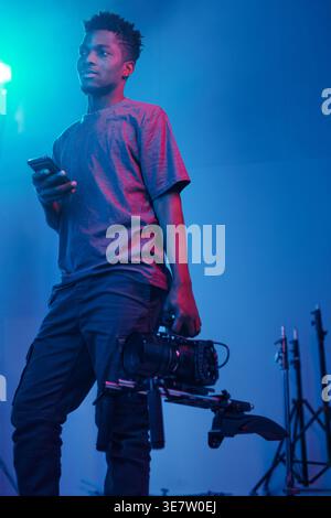 Cropped image of Black cameraman using smartphone while standing with professional camera in studio Stock Photo