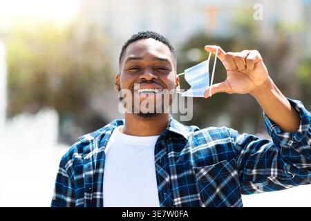 Smiling Black man taking off face mask standing on street on sunny day Stock Photo