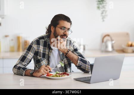 Arab man wearing eyeglasses having snack and using laptop while sitting at table in kitchen at home Stock Photo