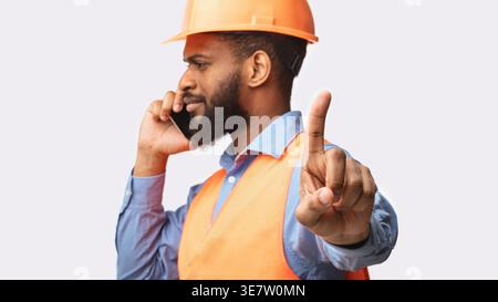 Black male worker wearing safety helmet having phone call and pointing finger forward looking away while standing against white background in studio Stock Photo