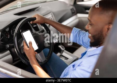 Smiling Black man wearing blue long sleeves using smartphone while sitting on driver seat holding steering wheel in car Stock Photo