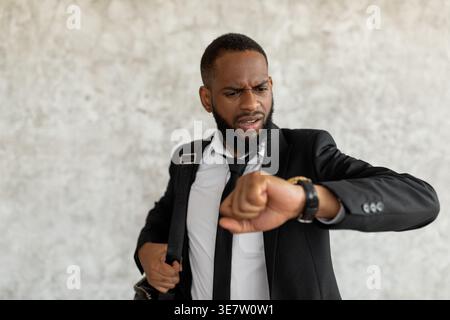 Worried Black businessman with open mouth looking at watch standing against gray wall Stock Photo