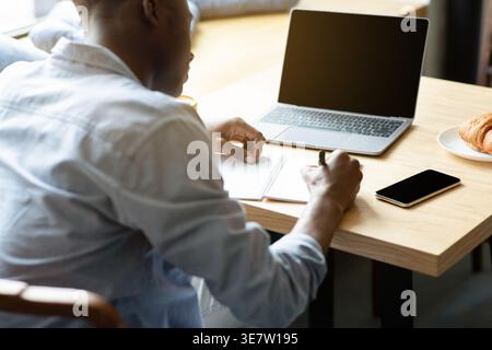 Cropped image of Black male student writing on notebook sitting in front of laptop at table in coffee shop Stock Photo