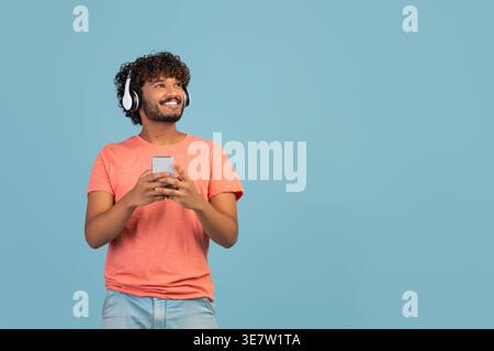 Happy millennial eastern guy in pink t-shirt using brand new wireless stereo headset and smartphone, listening to music over blue studio background, l Stock Photo