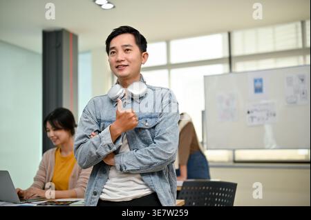 Smiling Asian male office worker wearing headphones around neck showing thumbs up and looking away while standing with crossed arms near colleagues... Stock Photo