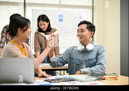 Smiling Asian male office worker giving high five to female coworker while sitting at table near colleagues working together in office Stock Photo