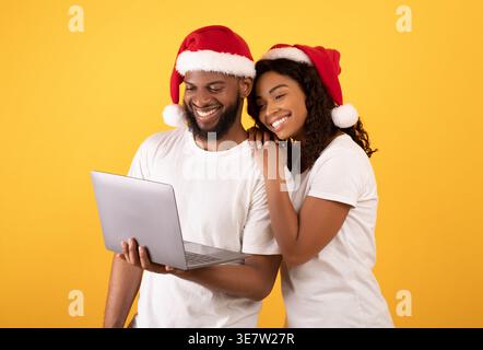 Smiling Black couple wearing Santa hats holding and looking at laptop standing against yellow background in studio Stock Photo