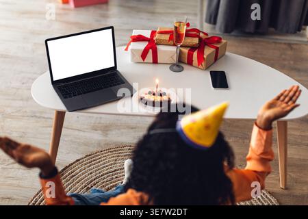 Black woman with curly wearing party hat raising opened hands while sitting at table with laptop and cake in living room at home Stock Photo