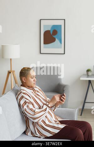 Vertical side view portrait of modern mixed raced woman using smartphone while sitting on sofa in minimal home interior Stock Photo