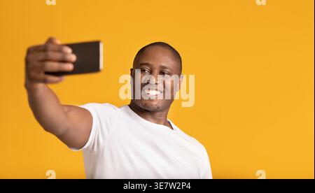 Smiling Black man in white shirt taking selfie using smartphone while standing against orange background in studio Stock Photo