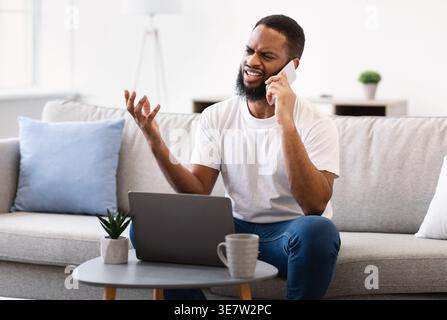 Black man with laptop talking on smartphone while sitting on sofa in living room at home Stock Photo