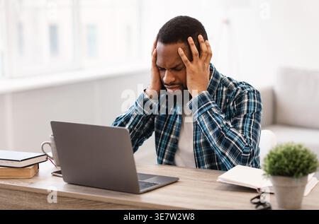 Stressed Black businessman having headache while sitting at table with laptop in living room at home Stock Photo