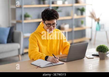 Smiling Asian man wearing eyeglasses and yellow hoodie taking notes and using laptop sitting at desk at home Stock Photo