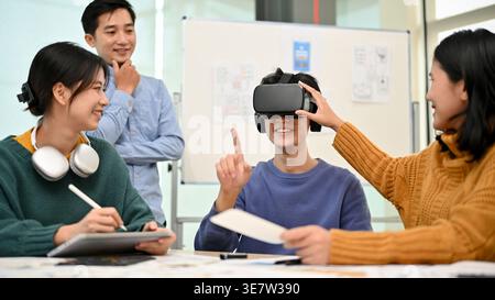 Smiling Asian male developer wearing VR headset sitting at table and testing newly developed VR game with colleagues in office Stock Photo