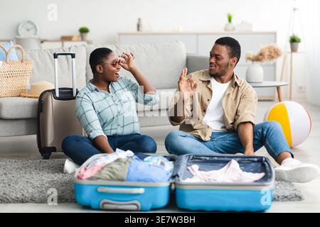 Cheerful Black man gesturing approving wifeâ€™s sunglasses while sitting on floor with packed suitcase in living room at home Stock Photo