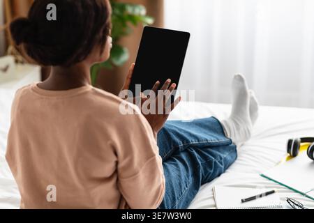 Black woman using tablet sitting on bed with notebooks in bedroom at home Stock Photo