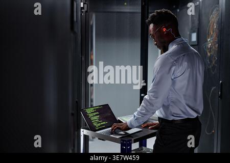 Black male network engineer using laptop and setting up servers while standing in data center Stock Photo