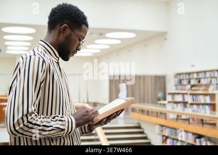 Black man wearing stripe long sleeves reading book while standing in library Stock Photo