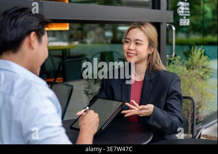 Thai businesswoman gesturing hand showing tablet to male client sitting at table in coffee shop Stock Photo