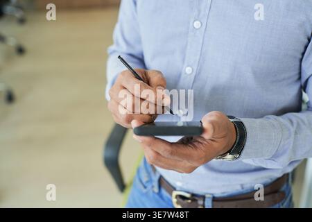 Hands of Indian male designer using smartphone with stylus pen while sitting on chair in office Stock Photo