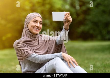 Arab woman wearing hijab taking selfie using smartphone while sitting in park during daytime Stock Photo