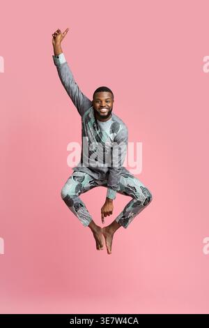 Excited black man in traditional grey clothes jumping up over pink studio background, full length photo. Joyful african american guy having fun while Stock Photo