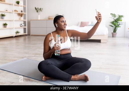 Curvy Black woman in wireless earphones holding bottle of water taking selfie using smartphone while sitting on yoga mat in living room at home Stock Photo
