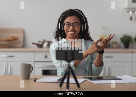 Black female tutor wearing eyeglasses and headphones recording video tutorial using smartphone gesturing hands while sitting at table with notebook... Stock Photo