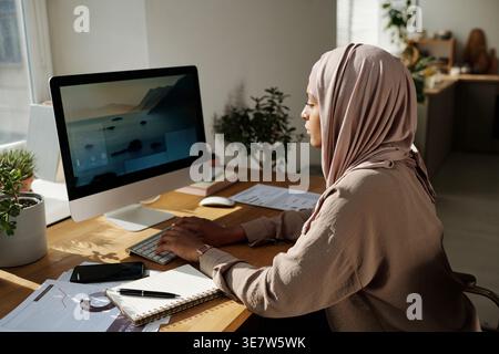 Black female programmer in hijab working on computer while sitting at table with smartphone and notepad at home office Stock Photo