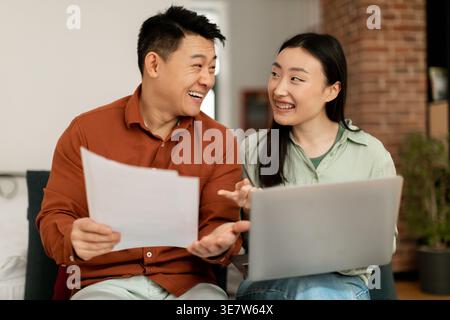 Cheerful Asian couple holding documents and laptop looking at each other while sitting on sofa in living room at home Stock Photo