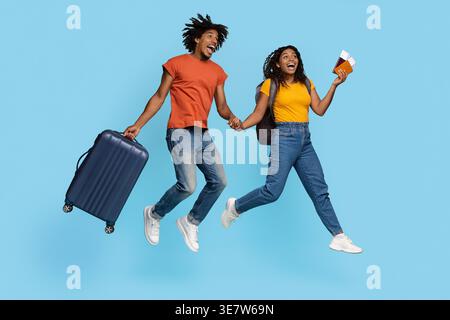 Cheerful Black man with open mouth carrying suitcase holding hands with girlfriend holding passports and tickets while jumping against blue backgro... Stock Photo