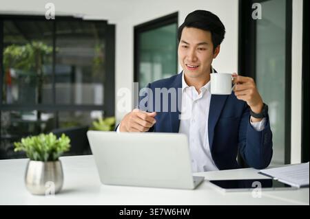 Asian businessman having online meeting on laptop holding coffee cup sitting at table in office Stock Photo