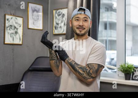 Smiling Asian male tattoo artist wearing cap looking away while standing and putting on latex gloves preparing for tattoo session in studio Stock Photo
